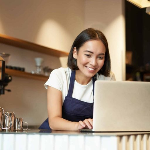 Portrait of smiling asian barista, cafe owner entrepreneur, working on laptop, processing orders on computer, standing behind counter.